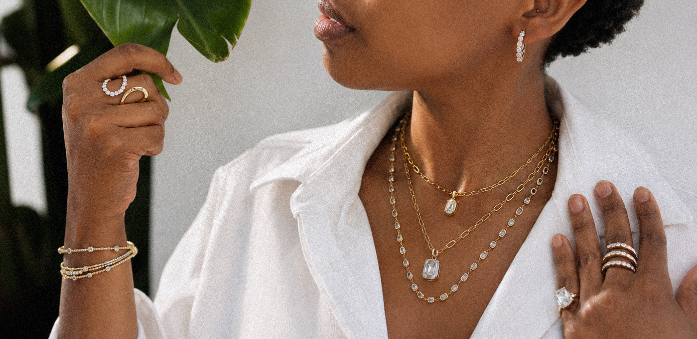 A close-up lifestyle shot of a woman wearing layered gold necklaces, bracelets, and rings, styled with a white collared shirt and holding a green leaf.